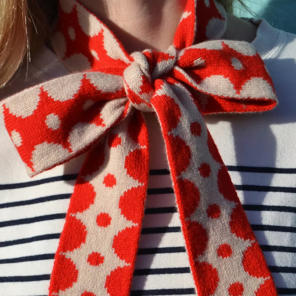 Close up of a model wearing a Scarlet Knitwear brand skinny 100% lambswool scarf, tied in a bow. It has a red and cream geometric design.