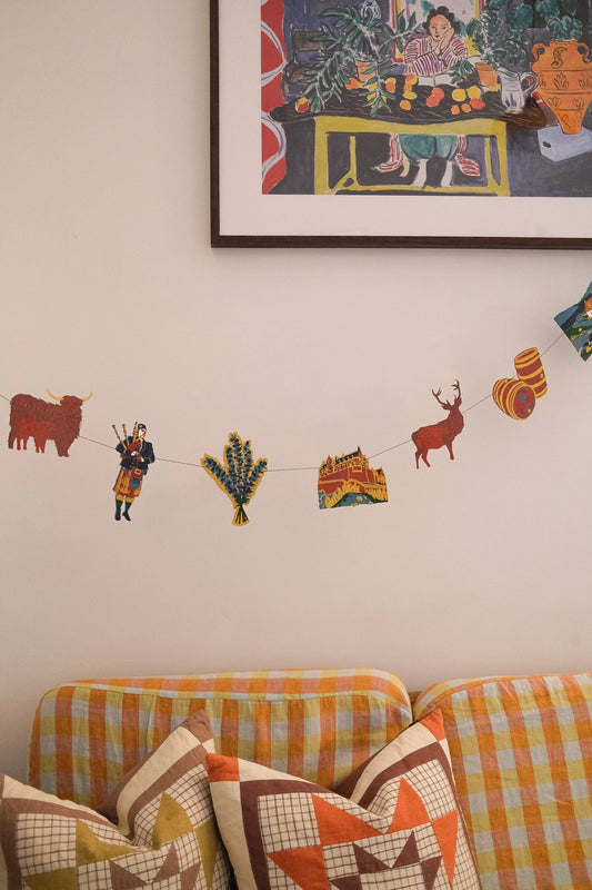 A colourful hand printed paper garland of various Scottish motifs, hanging above a sofa.