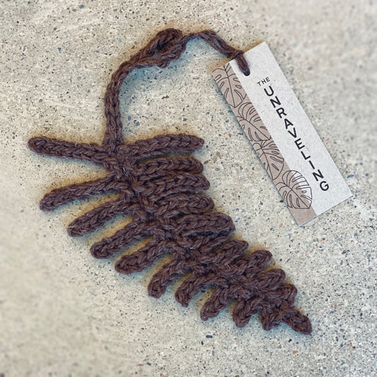 A decorative fern ornament, handmade from Library Brown Iona Wool with The Unraveling branded tag.
