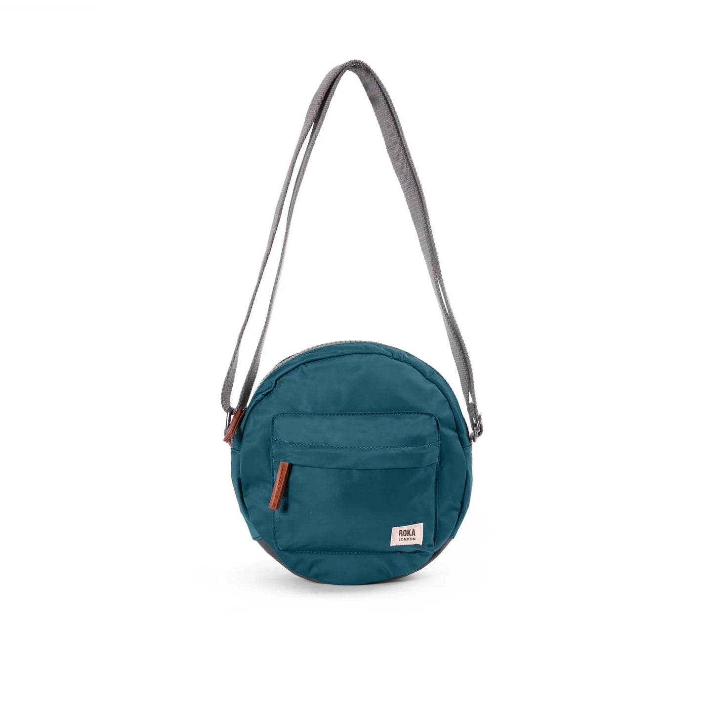 A small round crossbody bag in Teal, a bright lilac purple, with front zipped pocket, ROKA brand label and grey adjustable strap.