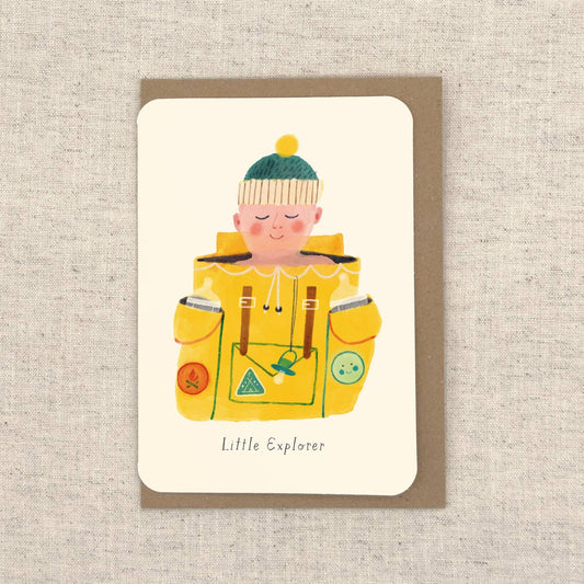 A greeting card with kraft envelope, featuring a colourful illustration of child wearing a wooly pompom hat sitting inside a yellow backpack and the words 'Little Explorer'.