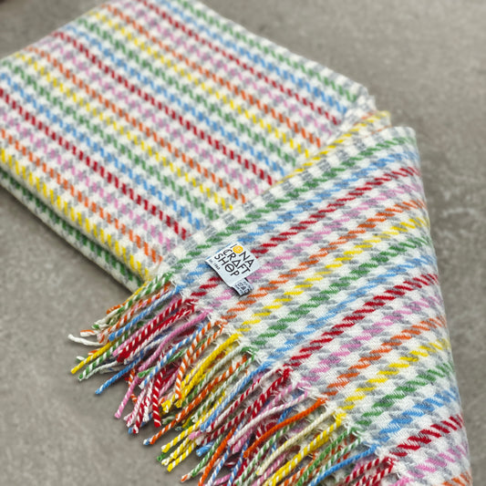 A folded 100% pure new wool blanket with a rainbow houndstooth pattern, fringed edge and Iona Craft Shop tag.