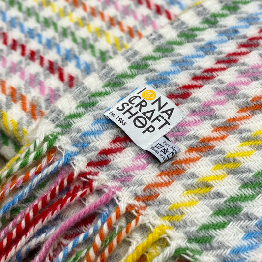 Close up of a folded 100% pure new wool blanket with a rainbow houndstooth pattern, fringed edge and Iona Craft Shop tag.