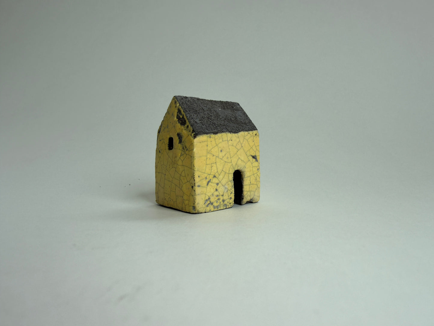 A small yellow ceramic miniature house with a dark gray glazed roof, photographed on a neutral light gray background.
