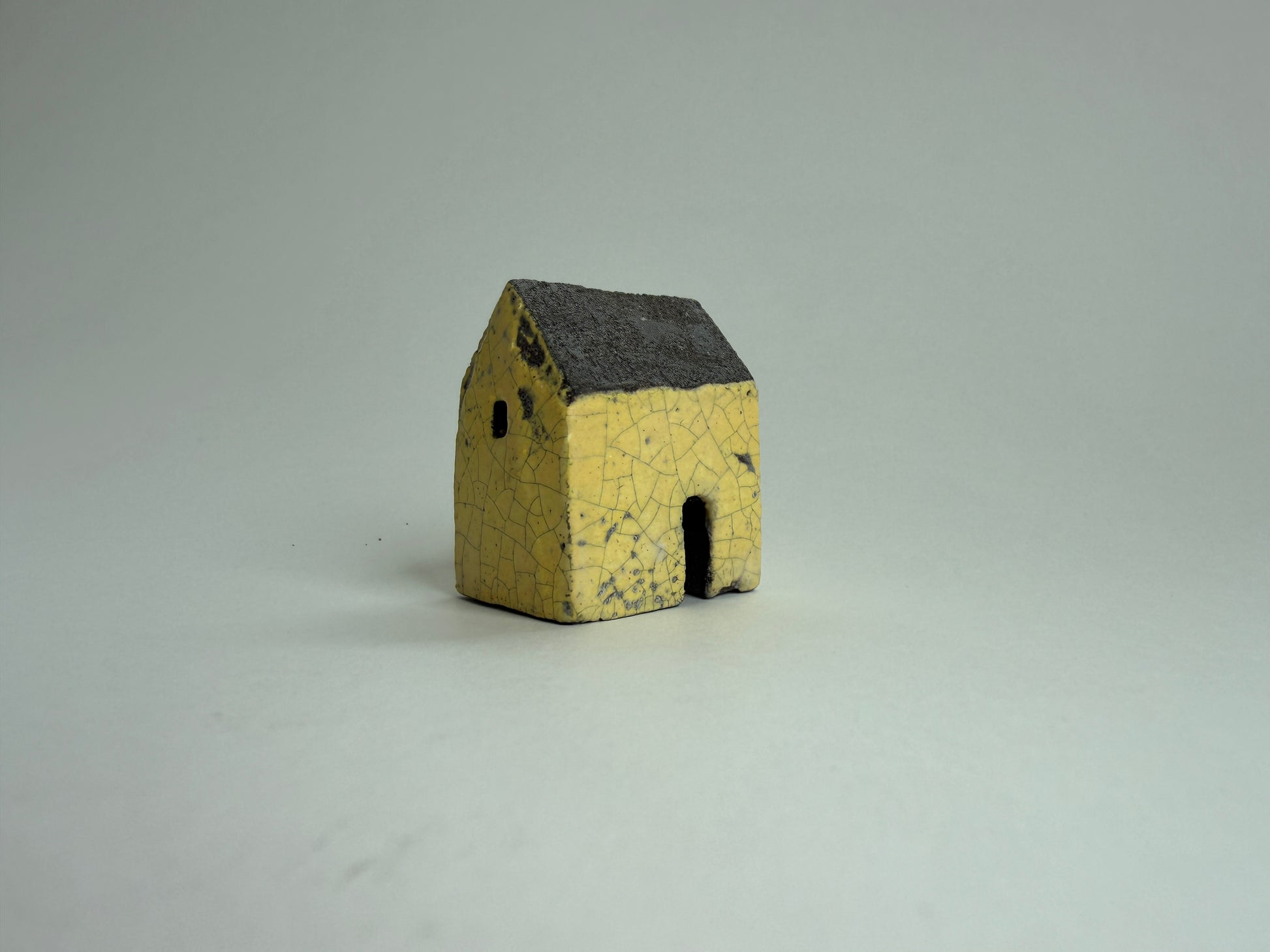 A small yellow ceramic miniature house with a dark gray glazed roof, photographed on a neutral light gray background.