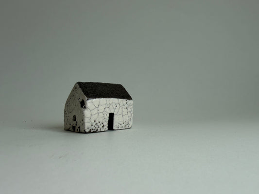 A small ceramic miniature house with a white body featuring a black crackle glaze pattern and a dark charcoal-colored roof, standing on a neutral light gray surface.