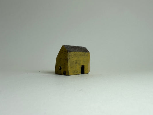 A small rustic ceramic house with a dark roof and weathered yellow-brown body, standing on a plain light gray background.