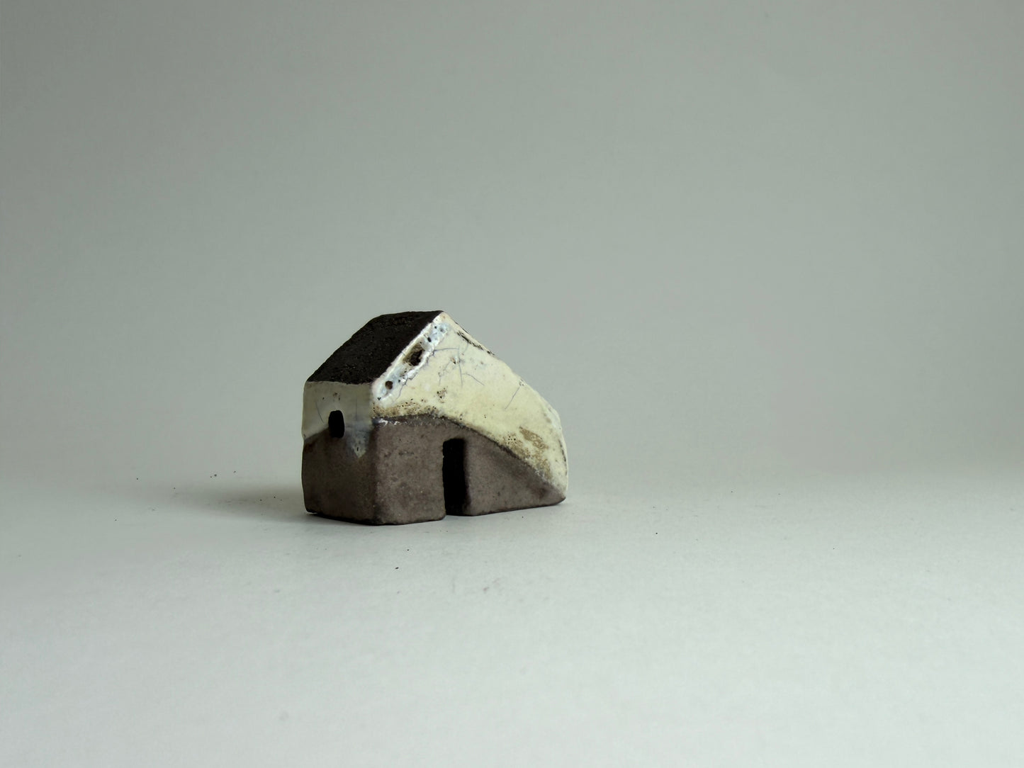 A small handmade ceramic house figurine with a dark brown base and a cream-colored roof, set against a plain light gray background.