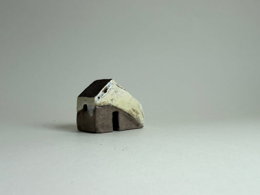A small handmade ceramic house figurine with a dark brown base and a cream-colored roof, set against a plain light gray background.