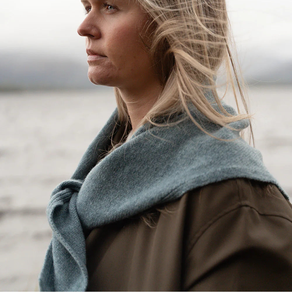 A woman in profile wearing a thick, soft blue-gray knit scarf and a brown jacket, standing outdoors 