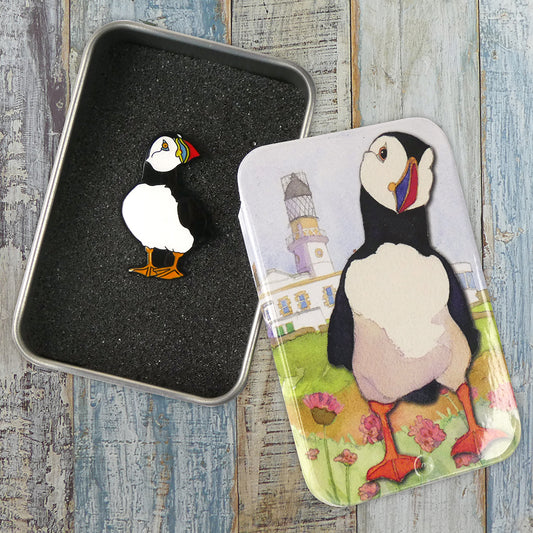 An enamel pin of a puffin, packaged inside a rectangular metal tin with an illustration of a puffin in front of a lighthouse with grass and thistles.
