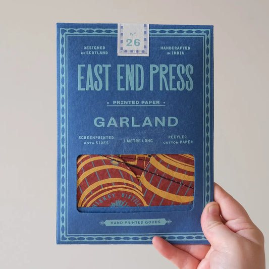 A hand holding a colourful hand printed paper garland with a Scottish theme, packaged in an East End Press branded envelope.