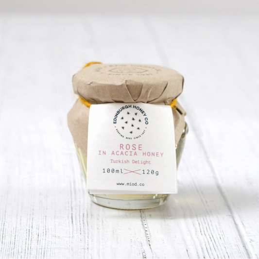 A small glass jar of Rose in Acacia Honey by The Honey Way Co., wrapped in brown kraft paper and a white paper label, set on a white wooden surface against a light gray background.